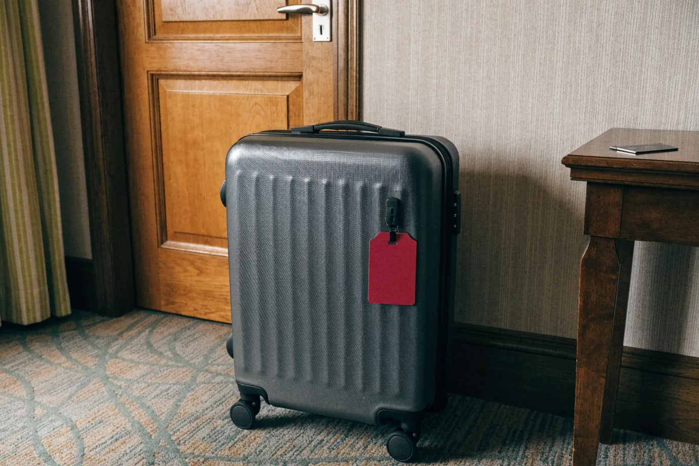 A carry-on suitcase by a hotel room door with a blank crimson luggage tag