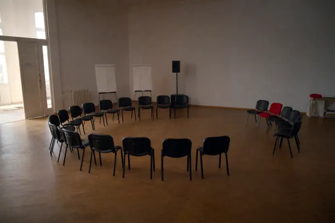 A circle of empty chairs in a bright workshop room, one of them crimson red