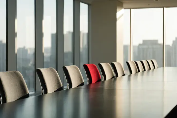 An empty boardroom with a row of grey chairs and one crimson chair by the window