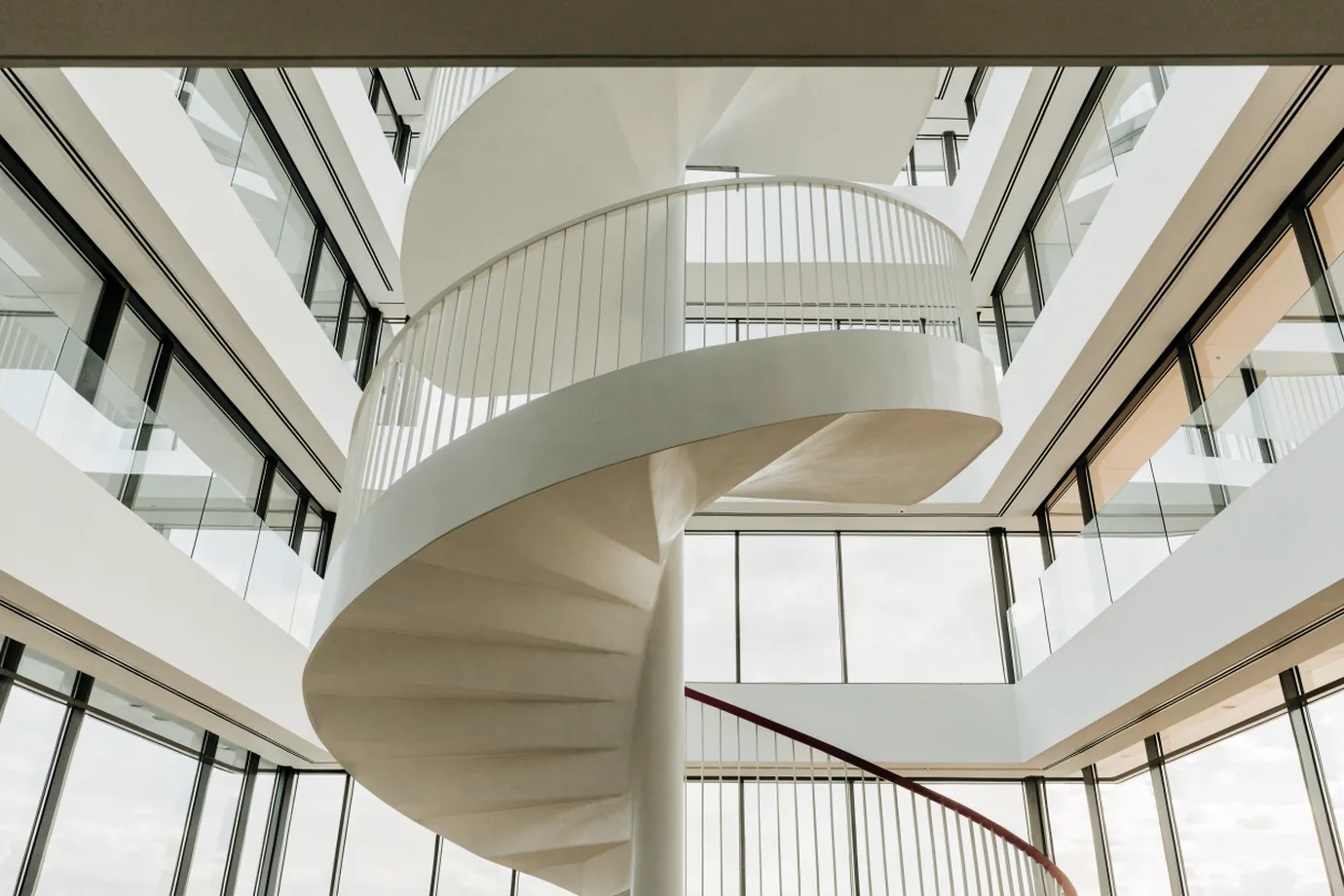 A white spiral staircase in an office atrium with a short crimson section of handrail